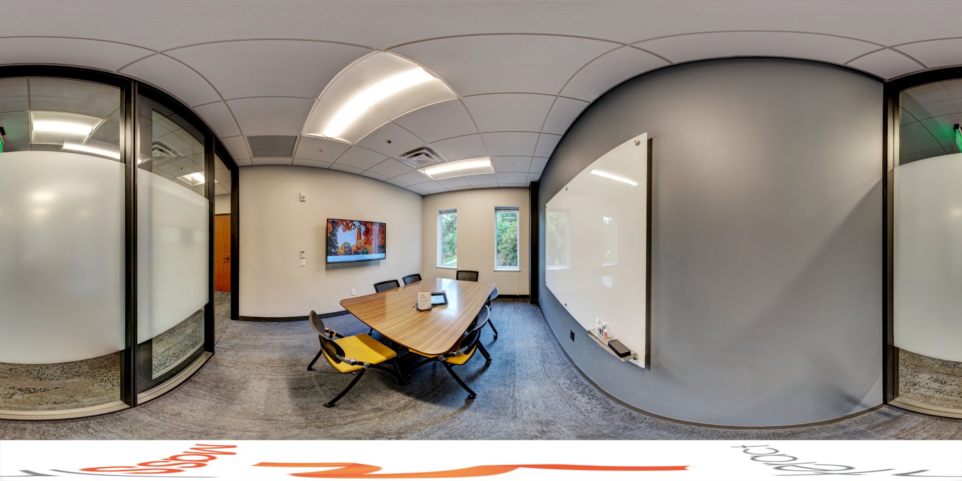 A panoramic view of a small meeting room with a rectangular table and chairs. The room features a large whiteboard on one wall and a mounted screen on another. Two windows provide natural light. Glass walls separate the space from the rest of the building.
